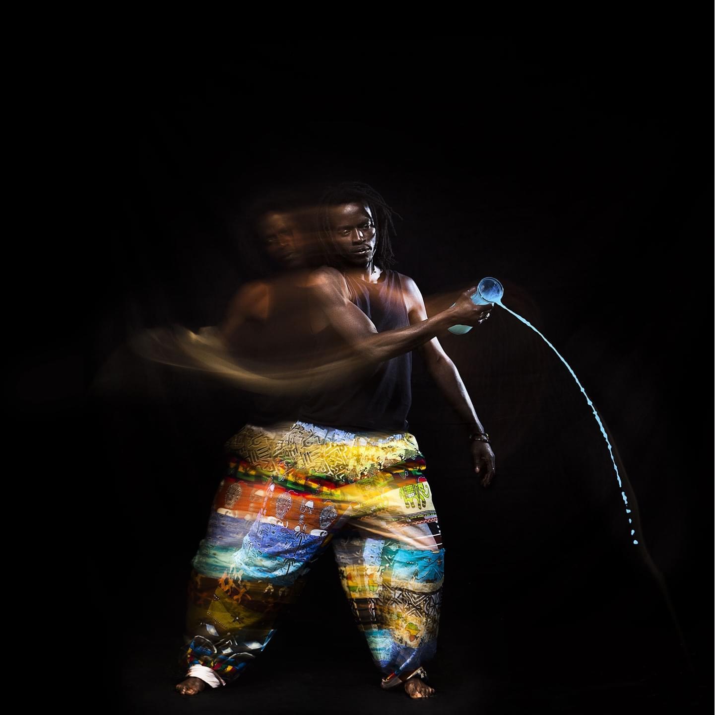 Senegalese dancer slowy moving his upper body from left to right while pouring stylized blue water out of a decanter.